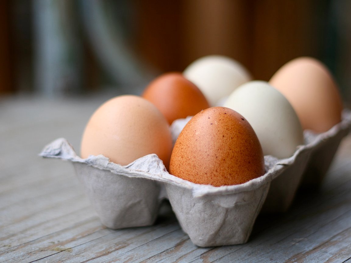 Fresh farm eggs in a carton featuring various colors, including brown, white, and cream, resting on a rustic wooden surface. Ideal for cooking and baking.